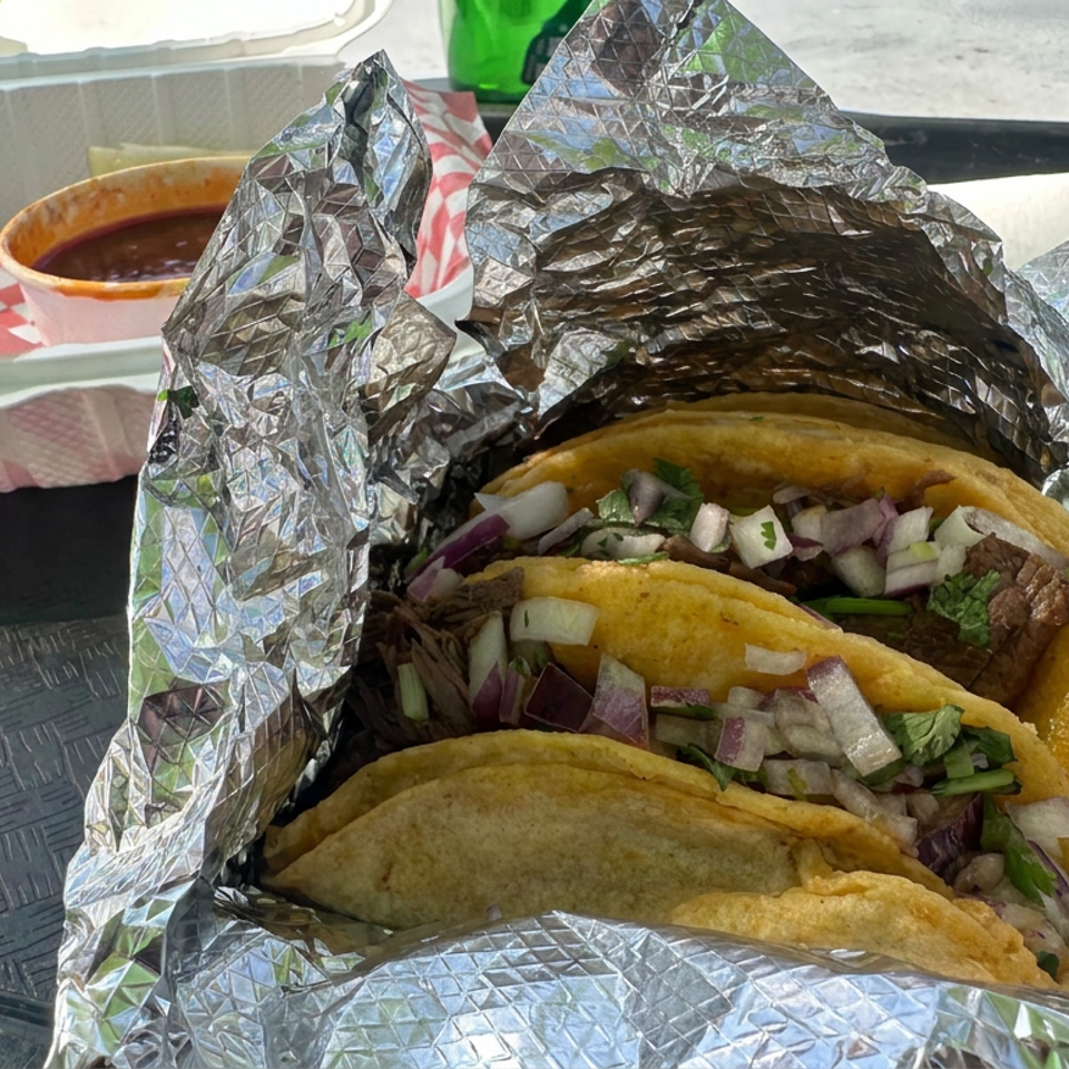 Three crispy tacos filled with meat, chopped onions, and cilantro sit in foil beside red salsa and a checkered paper-lined basket.